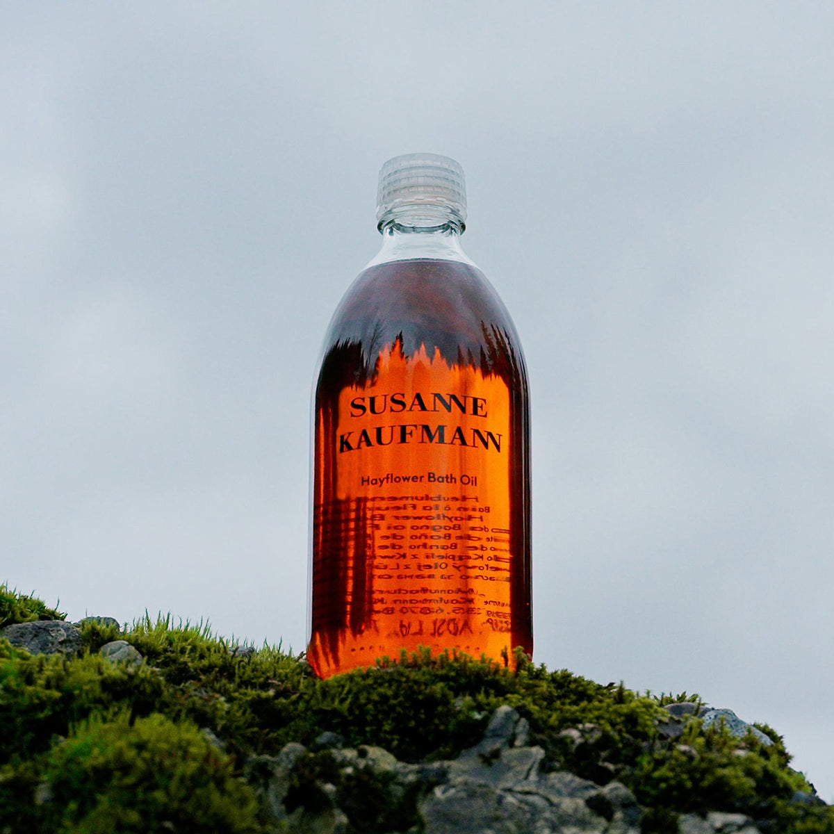 Susanne Kaufmann Hayflower Bath Oil / Heublumen-Ölbad, 250ml – Flasche auf Moos vor bewölktem Himmel