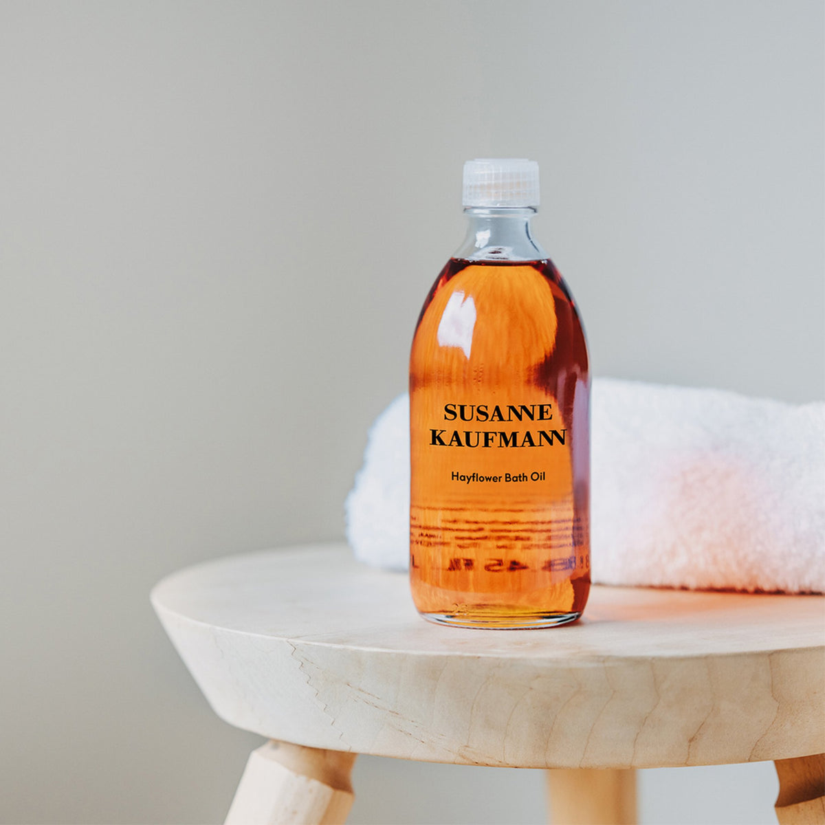 Susanne Kaufmann Hayflower Bath Oil / Heublumen-Ölbad, 250ml – Flasche auf einem Holzhocker mit Handtuch